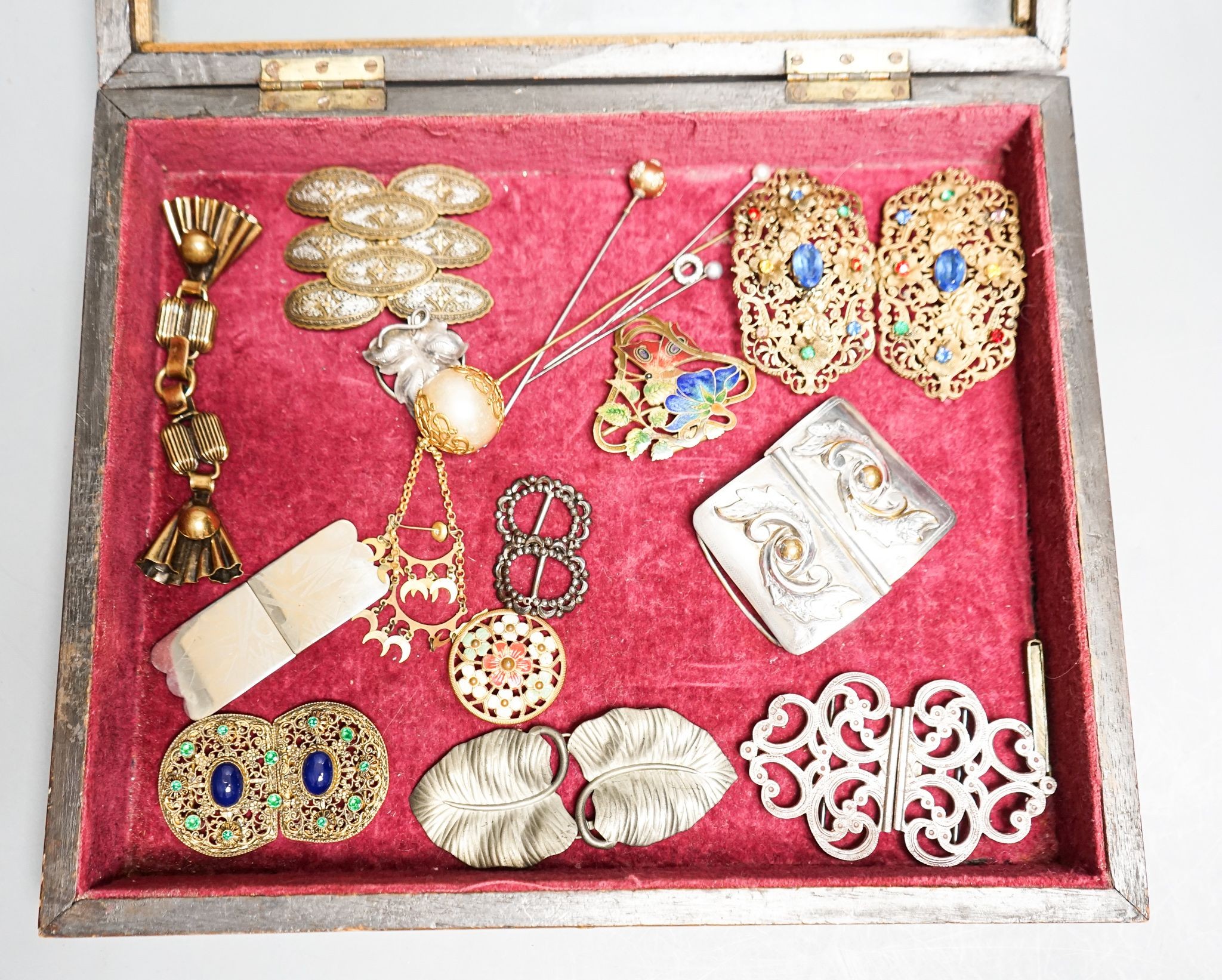 A group of belt buckles, clips and hatpins in a mahogany display case, 30.5cm